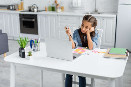 Bored schoolkid holding pen near notebooks and laptop in kitchenの写真素材
