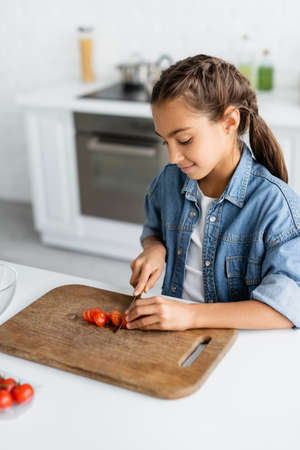 Smiling kid cutting cherry tomato on wooding cutting board in kitchenの写真素材