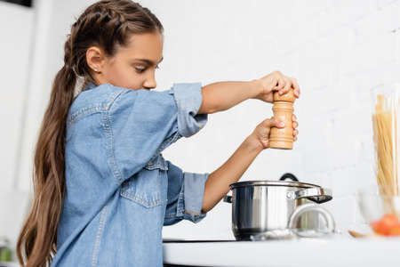 Preteen kid holding salt mill near saucepan in kitchenの写真素材
