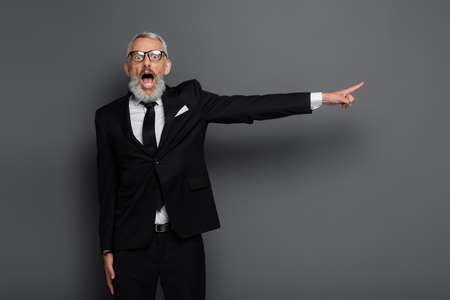 shocked middle aged businessman in glasses pointing with finger on gray