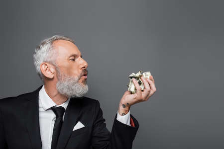 side view of bearded middle aged businessman blowing on crumpled dollar banknotes isolated on gray