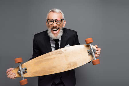 joyful and mature businessman in suit and glasses holding longboard isolated on grayの写真素材