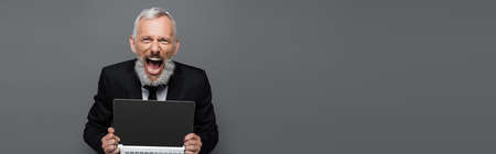 amazed middle aged businessman in suit holding laptop with blank screen on gray, bannerの写真素材