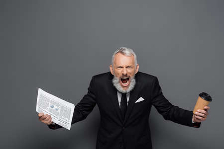 angry middle aged businessman holding paper cup and newspaper while screaming on gray
