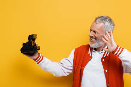 happy middle aged man in bomber jacket taking selfie on black vintage camera on yellowの写真素材