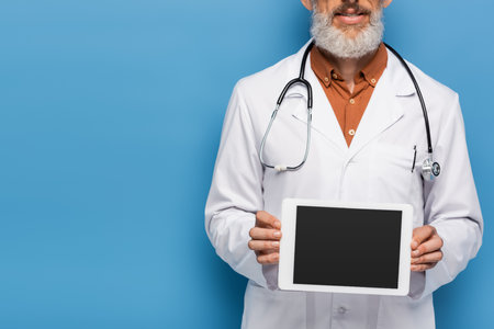 cropped view of bearded middle aged doctor in white coat holding digital tablet with blank screen on blueの写真素材
