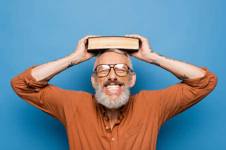 joyful middle aged teacher in glasses holding book on head on blueの写真素材
