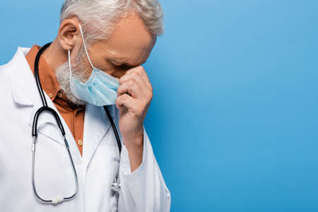 upset middle aged doctor in white coat and medical mask on blueの写真素材