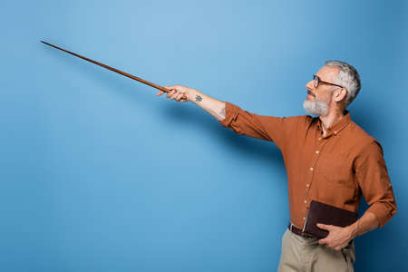 tattooed middle aged teacher in glasses holding pointer stick and book on blueの写真素材