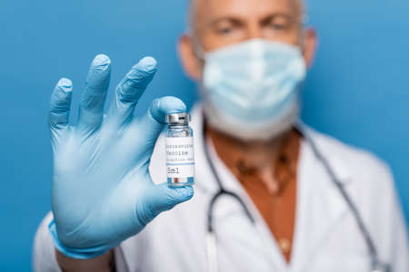 blurred doctor in medical mask holding vial with vaccine isolated on blueの写真素材