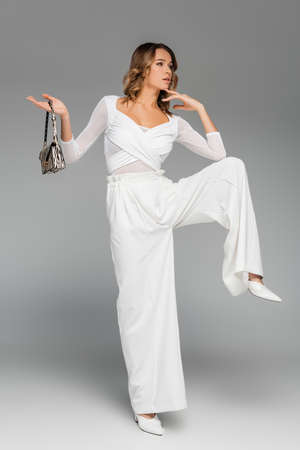 full length of curly young woman in white outfit holding purse with animal print and posing on grayの写真素材