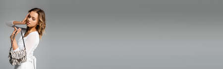 curly young woman in white outfit holding purse with animal print on gray, bannerの写真素材