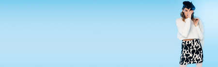 young woman in skirt with cow print holding white faux fur jacket while adjusting beret on blue, bannerの写真素材