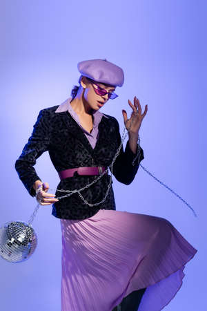 stylish woman in sunglasses and blazer with animal print holding disco ball on chain on violet