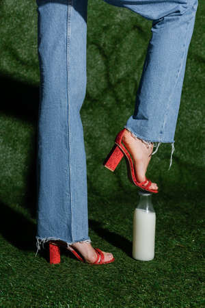cropped view of stylish model in red high heels and jeans stepping on glass bottle of milk near...