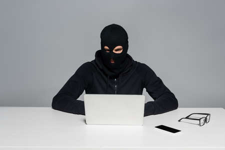 Hacker in balaclava using laptop near smartphone and eyeglasses on table isolated on grayの写真素材