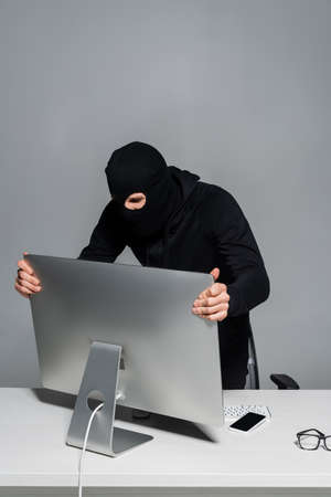 Hacker in balaclava taking computer monitor near smartphone isolated on gray