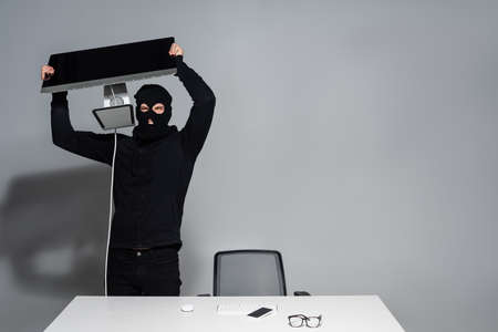 Hacker in balaclava holding computer monitor near cellphone on table on gray backgroundの写真素材