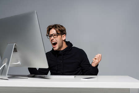 Angry programmer in eyeglasses screaming near computer and smartphone isolated on grayの写真素材