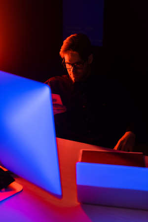 Programmer in eyeglasses using smartphone near computers on table on black background