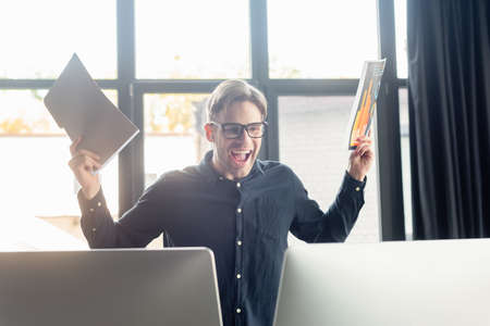 Excited programmer holding paper folder and document near computers in office