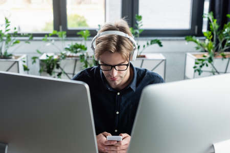 Young programmer in headphones using smartphone near computer monitorsの写真素材