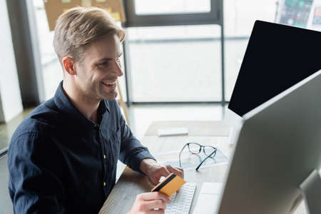 Cheerful programmer holding credit card while using computers in office