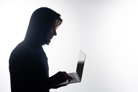 Side view of hacker in hood using laptop with blank screen isolated on grayの写真素材