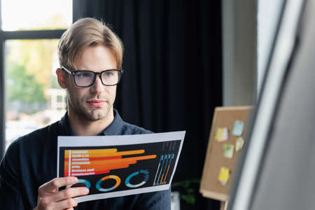 Young programmer holding document near blurred flip chart in officeの写真素材