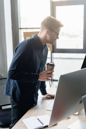 Side view of programmer in earphone holding paper cup near computers and notebook in officeの写真素材