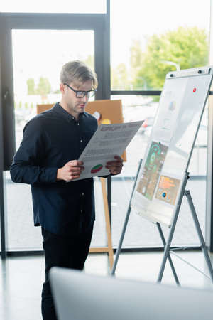 Programmer in eyeglasses holding document near flip chart and blurred computer in office