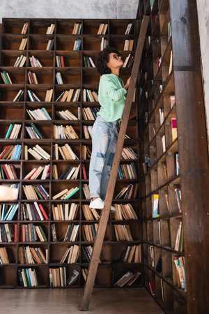 curly african american student standing on wooden ladder in libraryの写真素材