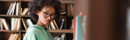 young african american woman in eyeglasses looking at camera near bookshelf, banner
