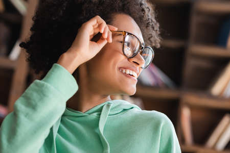 positive african american student smiling while adjusting eyeglasses and looking awayの写真素材
