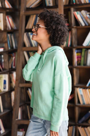 joyful african american student in eyeglasses looking at bookshelfの写真素材