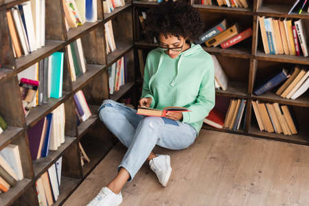 curly african american student in eyeglasses sitting on floor and reading book in libraryの写真素材