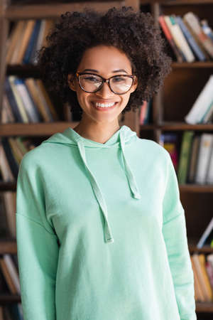 cheerful african american student in eyeglasses looking at camera near bookshelf