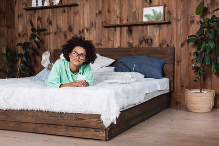 pensive african american woman in eyeglasses lying on bed while using smartphone near laptop
