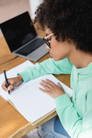curly african american student in eyeglasses writing in notebook while studying online near blurred laptop with blank screenの写真素材