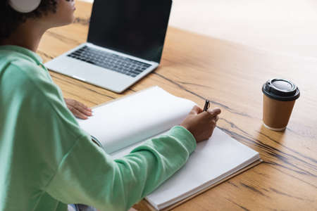cropped view of african american student writing in notebook near laptop with blank screen