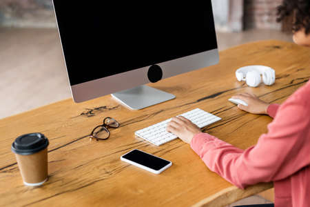 partial view of african american woman using computer keyboard near cellphone, paper cup and...