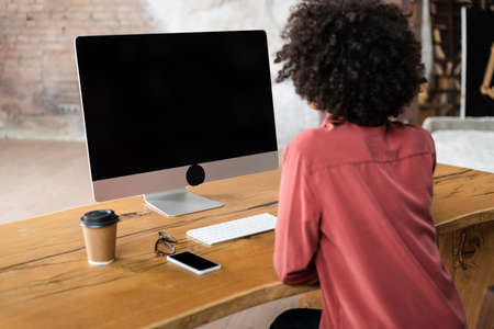back view of curly african american woman using computer near smartphone, paper cup and eyeglasses on deskの写真素材