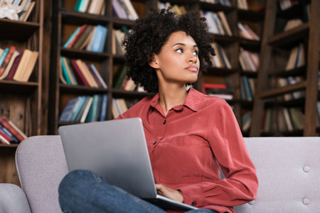 distracted african american woman sitting on couch with laptop and looking away