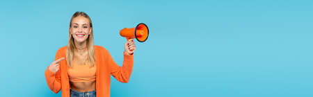 Smiling blonde woman pointing at loudspeaker isolated on blue, bannerの写真素材