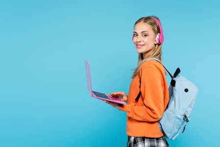 Blonde student in headphones and backpack holding laptop isolated on blue