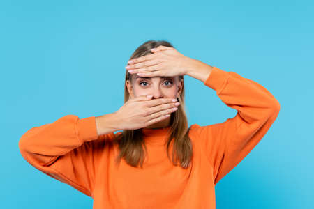 Blonde woman covering face and looking at camera isolated on blueの写真素材