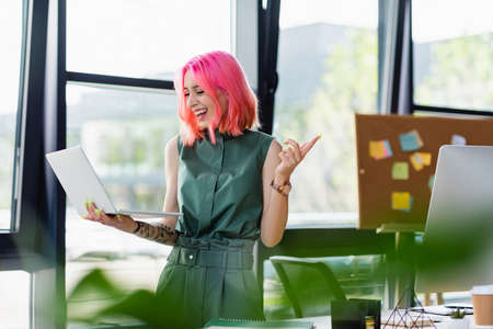 cheerful businesswoman with pink hair holding laptop and pointing with finger in officeの写真素材