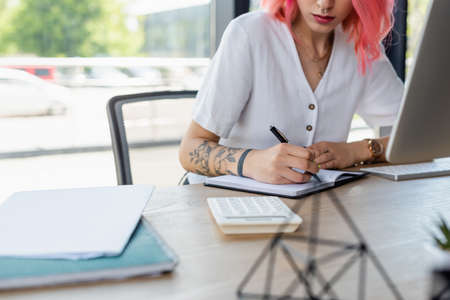 cropped view of pierced businesswoman with pink hair writing on notebook in officeの写真素材
