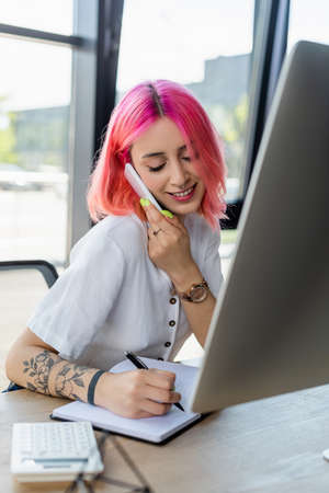smiling businesswoman with pink hair talking on smartphone and writing on notebook near monitorの写真素材