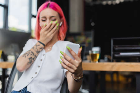blurred businesswoman with pink hair and earphone using smartphone and covering mouth while laughing...